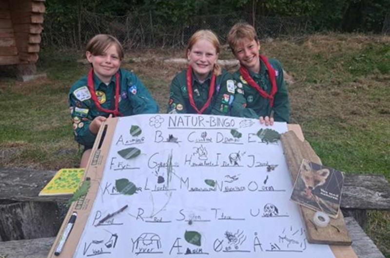 Tre spejdere der sidder med et stort stykke papir hvor der står "natur-bingo", og der er fundet ting fra naturen til hvert bogstav.