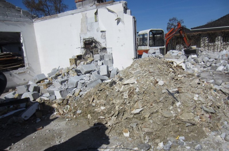 Hus under nedrivning. Foran huset ligger en bunke murbrokker. I baggrunden er en gravko.