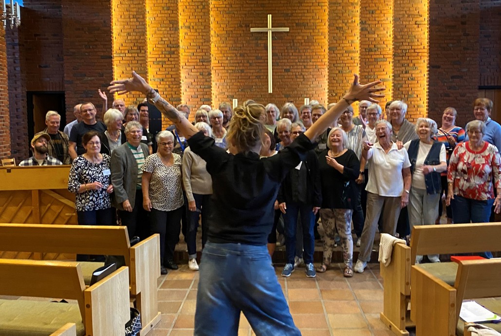 Kor der synger indenfor i en kirke. Dirigenten står energisk forrest med ryggen til kameraet. Der er et kors i midten.