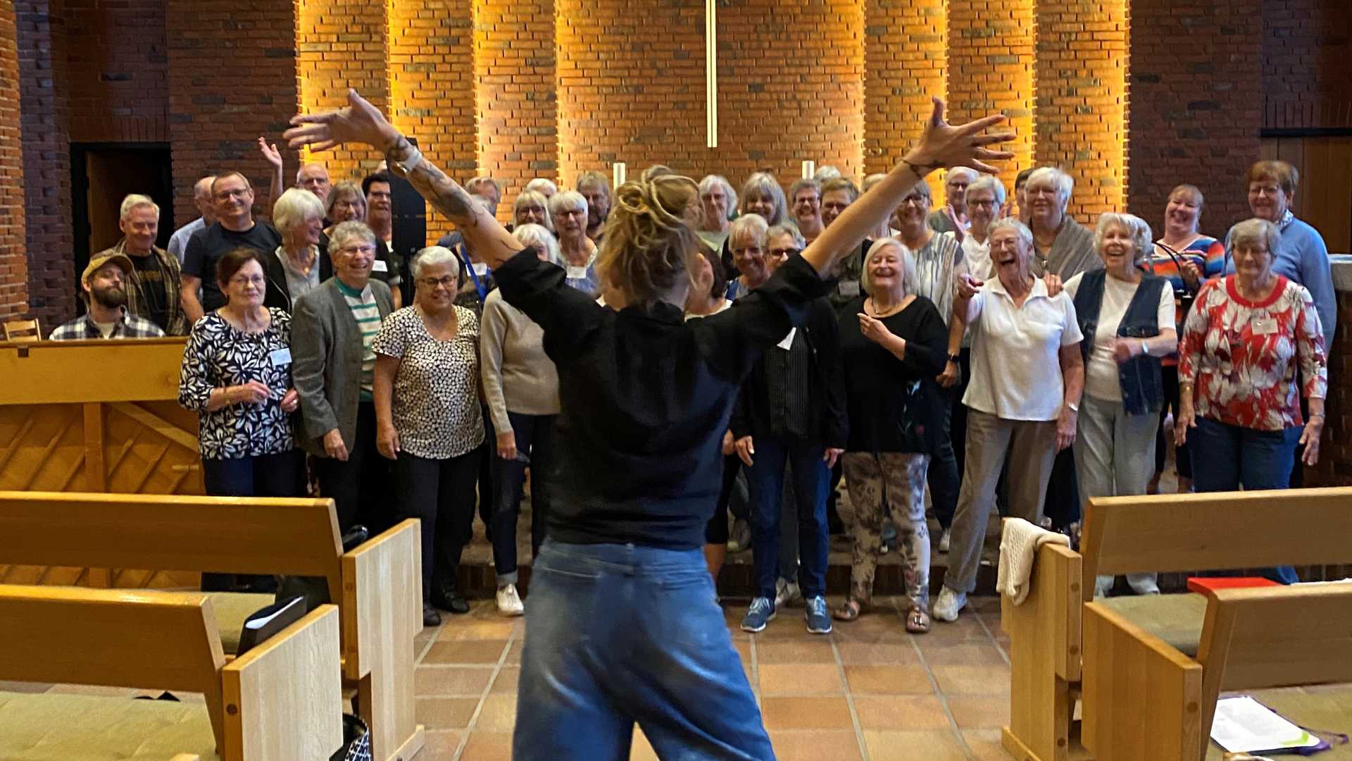 Kor der synger indenfor i en kirke. Dirigenten står energisk forrest med ryggen til kameraet. Der er et kors i midten.