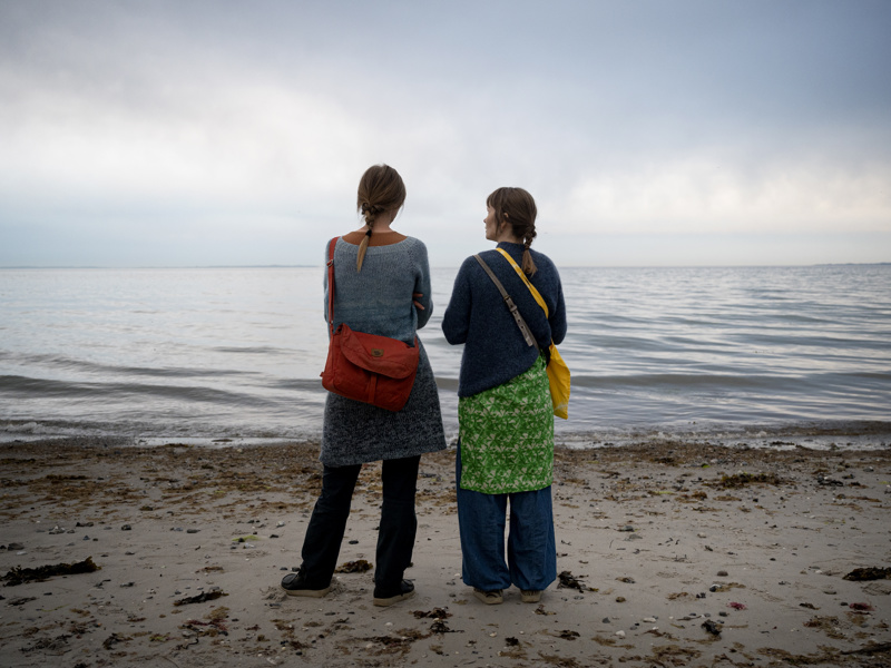 To kvinder står med ryggen til og kigger udover vandet på stranden