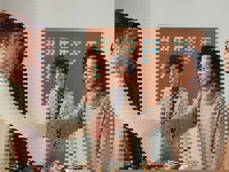 5 personer står og snakker sammen rundt om en bord, 2 af dem giver hinanden hånden over bordet