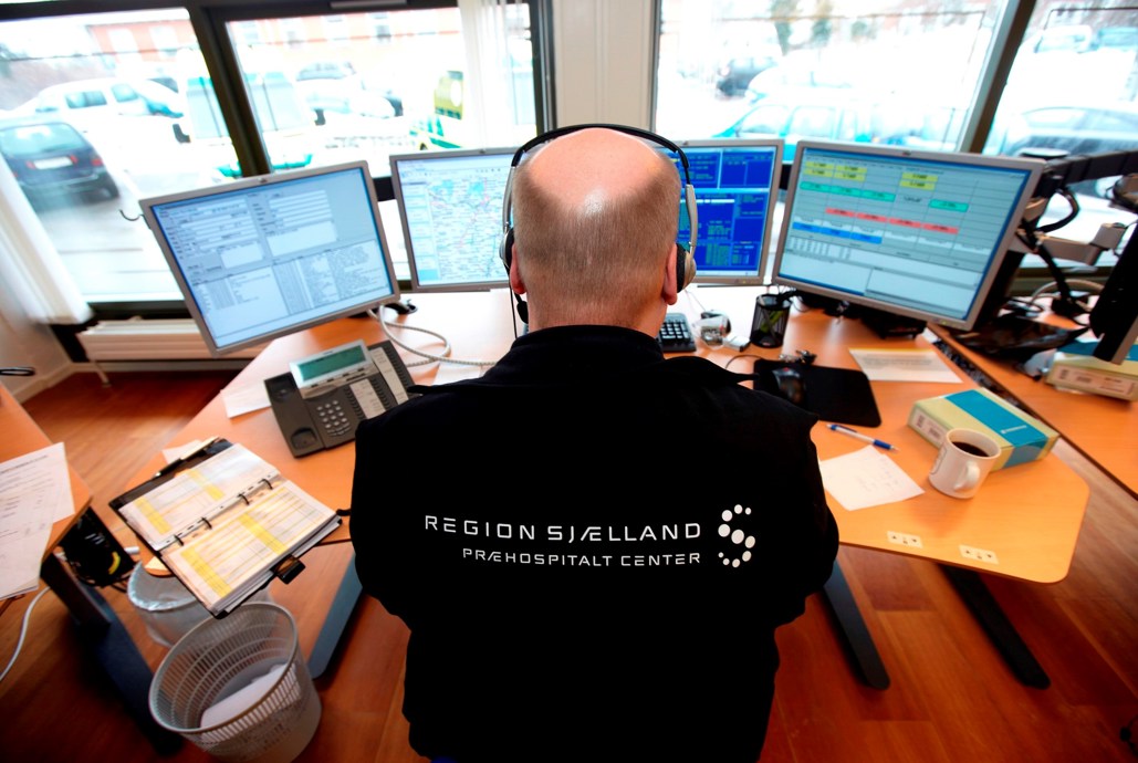 Medarbejder fra alarmcentralen sidder ved computerskærme. På ryggen af medarbejderen står der "Region Sjælland, Præhospitalt Center".