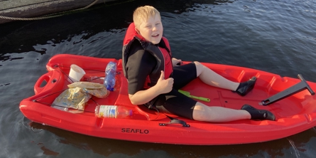 En dreng i en rød kajak på vandet der giver thumbs up og har samlet skrald der ligger bagerst i kajakken
