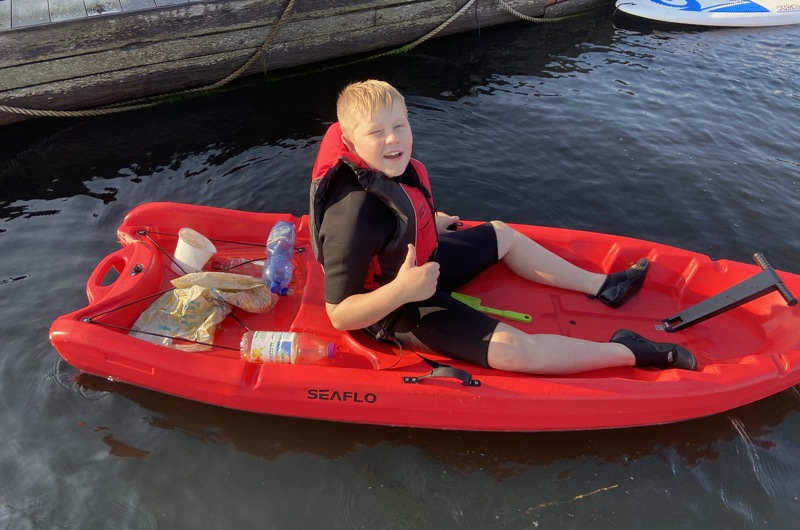 En dreng i en rød kajak på vandet der giver thumbs up og har samlet skrald der ligger bagerst i kajakken