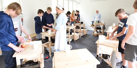 Elever der står med æbler ved træborde på Greve Museum, mens der går en dame rundt i udklædning