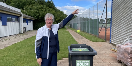 Ældre kvinde der står i tennisklubben og smider en tennisbold i en spand hvor der står "Kun brugte bolde - ingen affald".