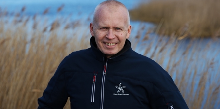 Mand der står og smiler foran en lille sø med siv i baggrunden