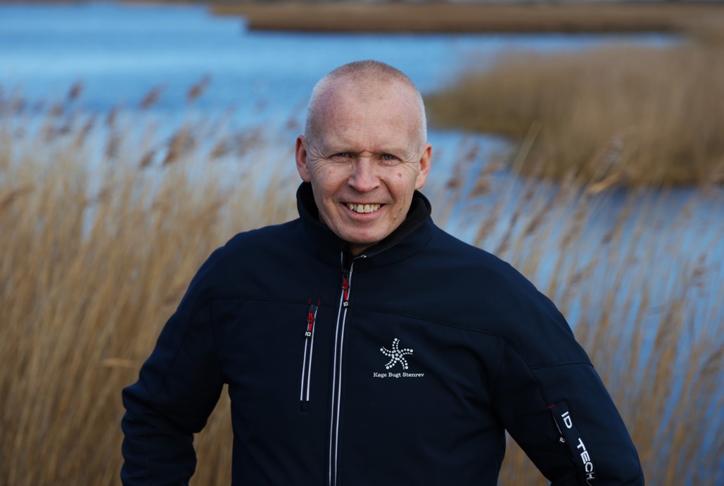 Mand der står og smiler foran en lille sø med siv i baggrunden