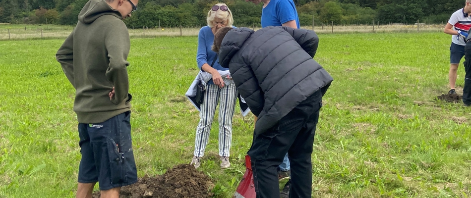 en underviser og tre elever der er udendørs og er i gang med at grave et hul i græsset, for at kunne plante mikroskoven. Det er godt vejr.