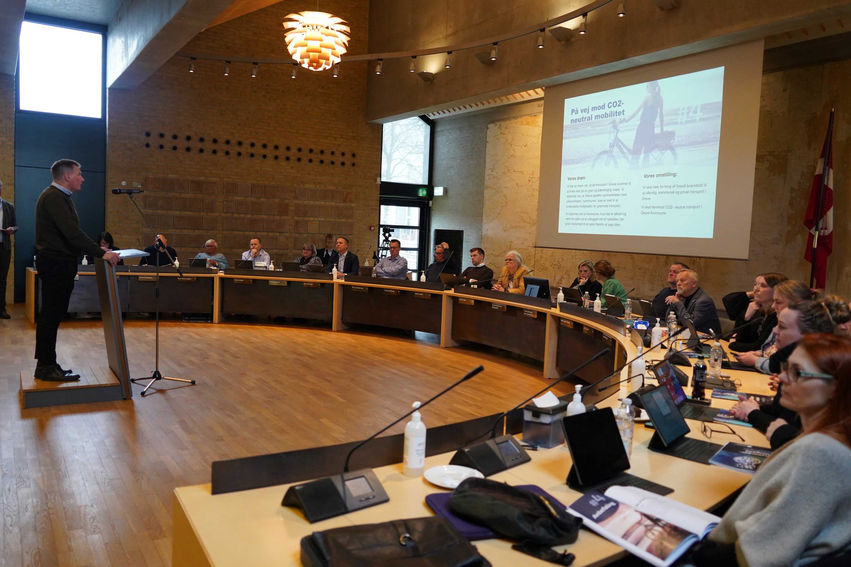 En mand præsenterer "På vej mod CO2-neutral mobilitet" mens der sidder folk og lytter i Greve Kommunes byrådssal.